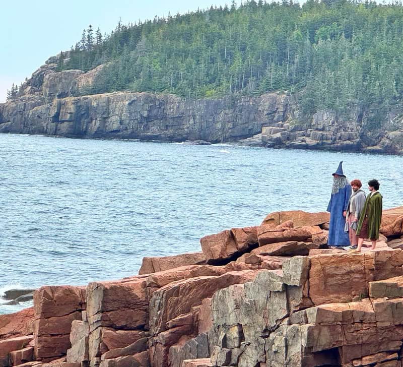 Three people in wizard costumes stand on rocks by the water with forested cliffs in the background.