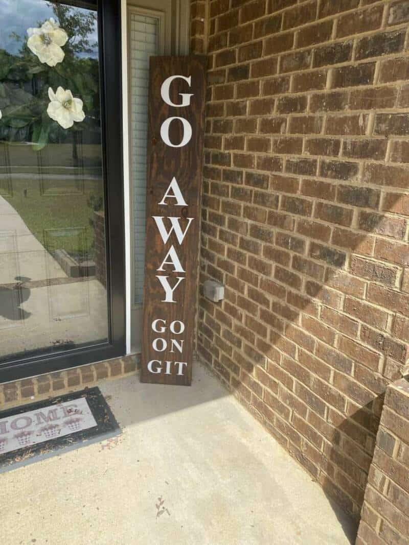 Wooden sign says 'GO AWAY GO ON GIT' next to a door on a brick wall.