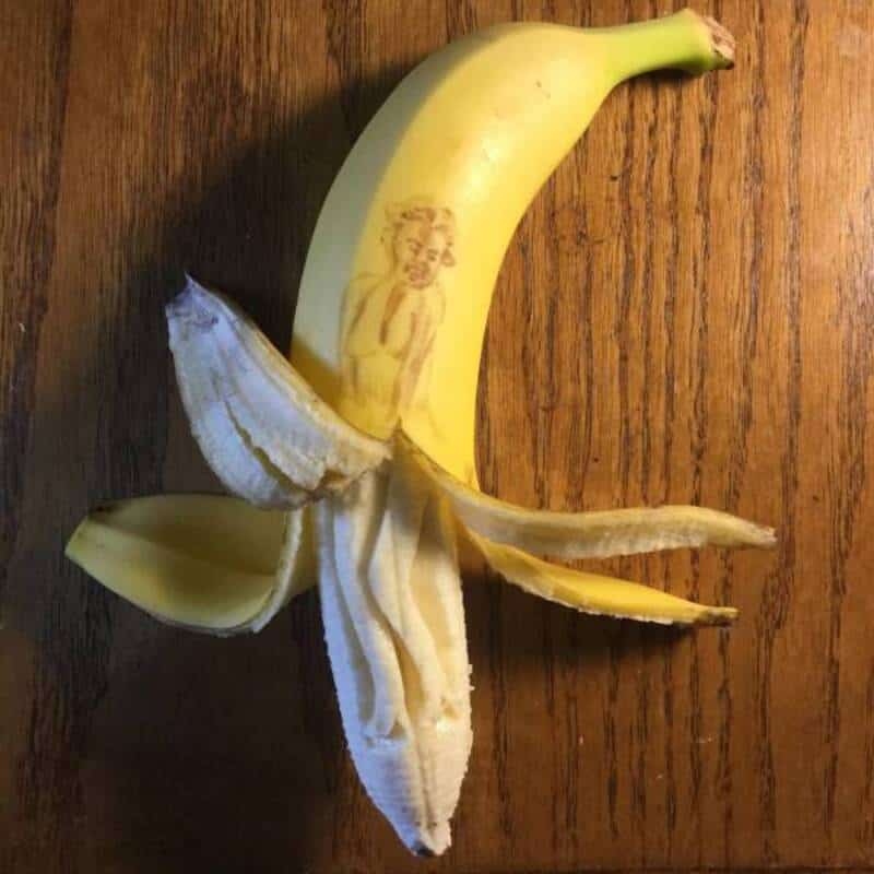Banana with a printed image of a woman on its peel, partially peeled on a wooden surface.