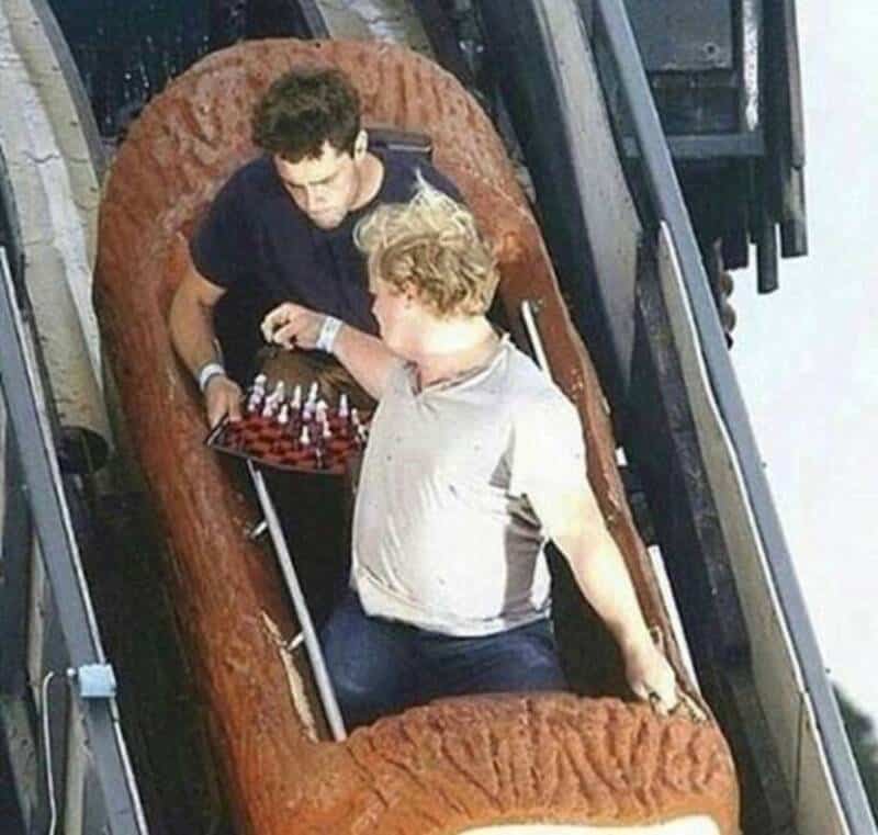 Two young men on a roller coaster, one holding a chessboard, with a humorous expression.