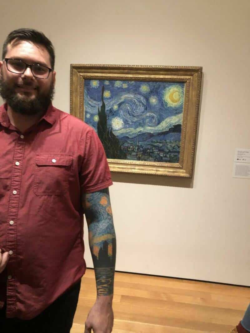 Man with glasses and beard standing in front of Van Gogh's Starry Night painting.