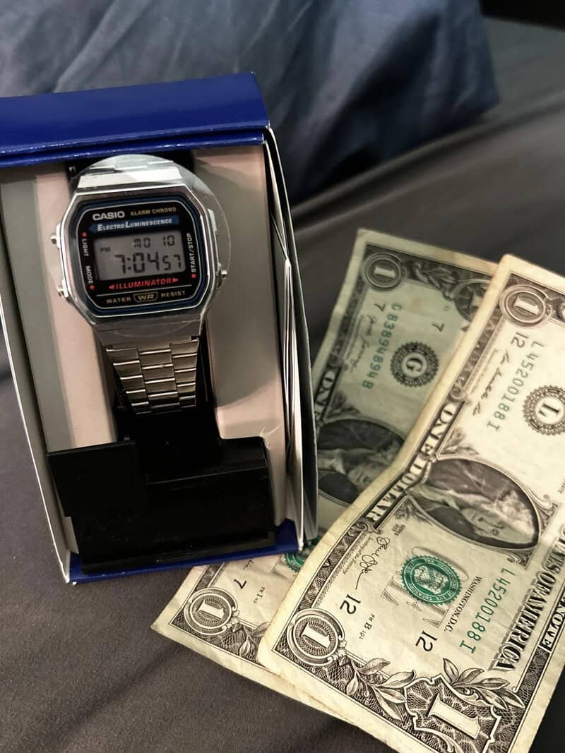 Casio digital watch in box with dollar bills nearby, on a dark surface.