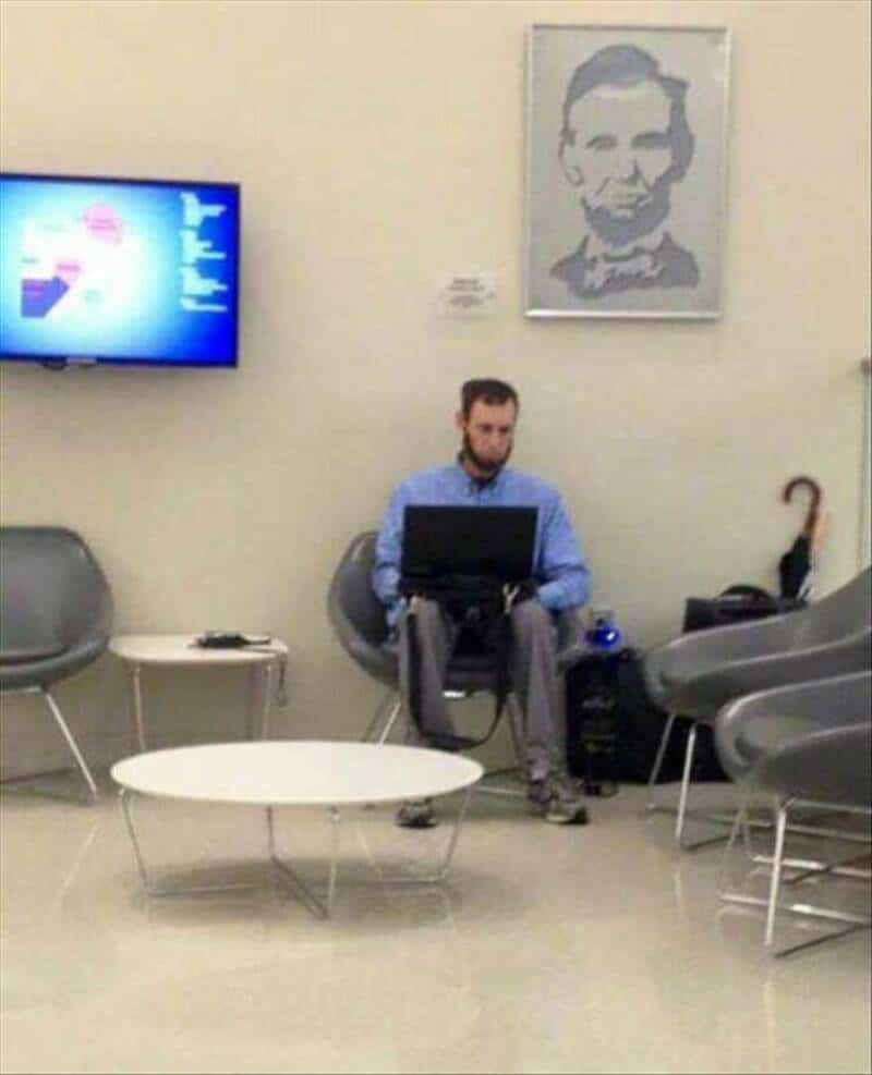 Man sitting alone in a waiting room with a laptop, portrait on the wall, TV on the wall.