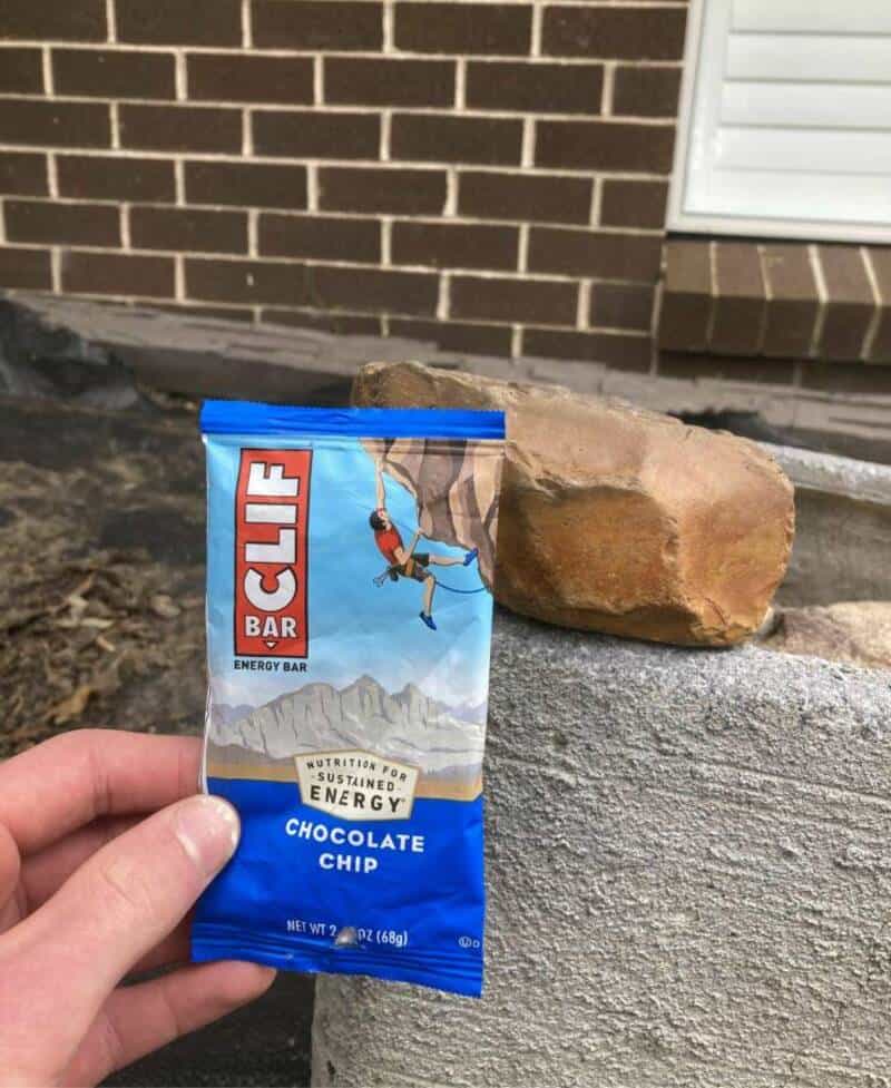Energy bar held in front of a large rock on a ledge outside.