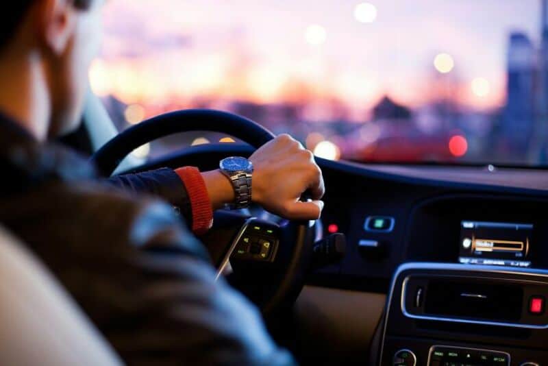 Person driving a car at sunset, city lights visible outside.