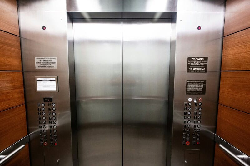 Elevator doors with control panels and warning signs on wood-paneled walls.