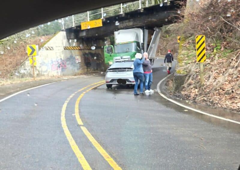 Car crashed into bridge support, causing damage, with three people nearby on a wet road.