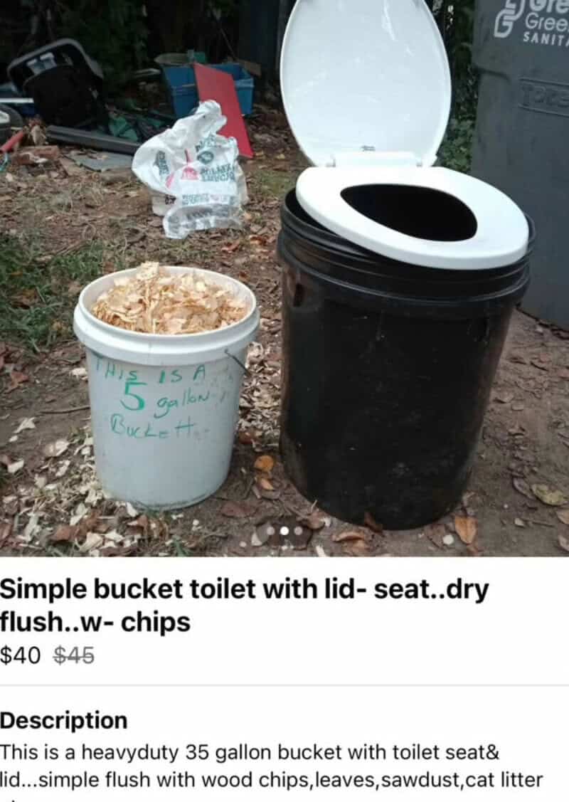 A bucket toilet with a lid, seat, and chips in a natural outdoor setting.