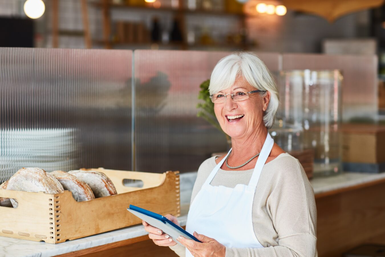 Smiling elderly woman in a bakery holding a tablet.