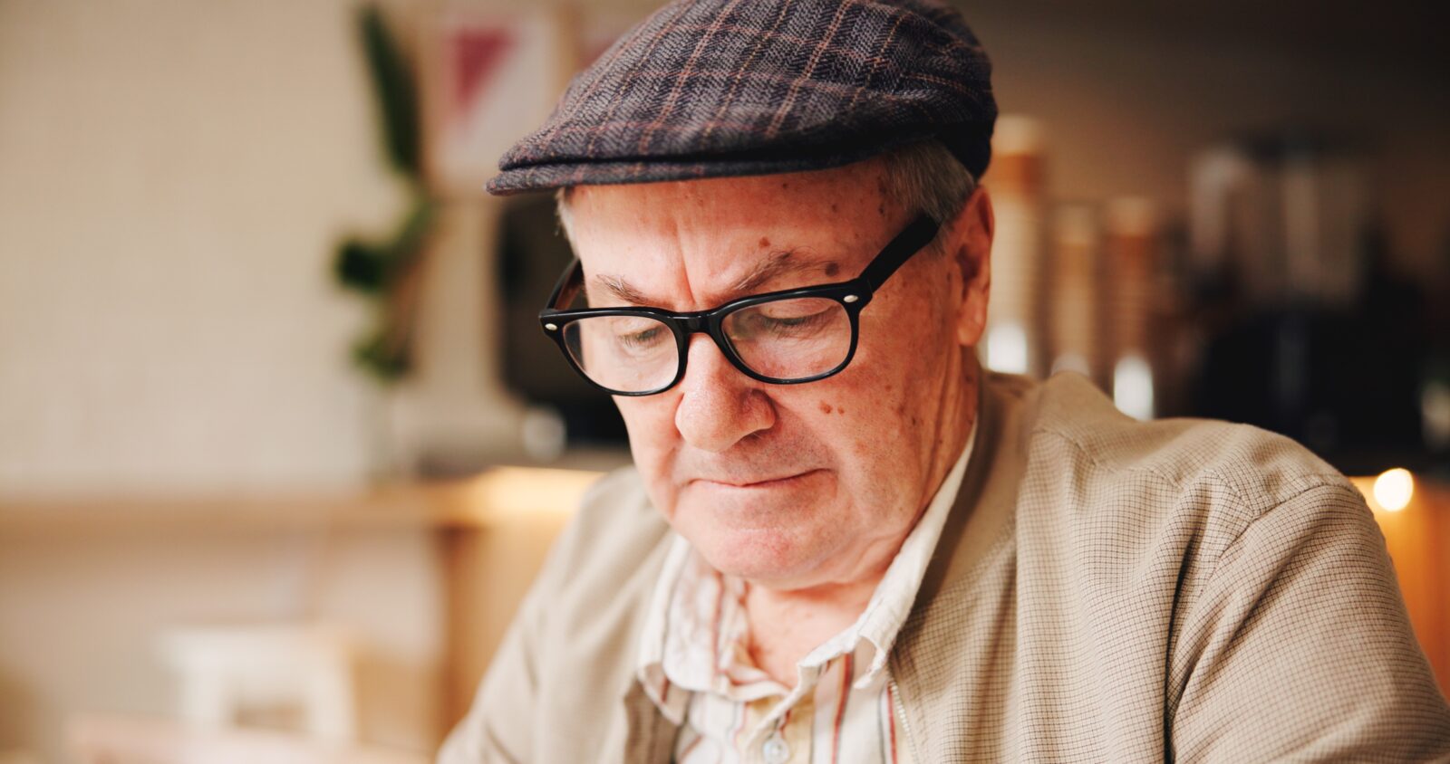 A man with glasses and a flat cap looking down, in an indoor setting.