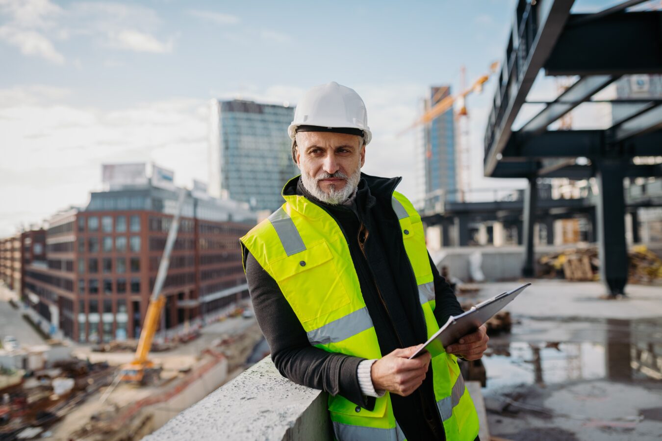 Construction worker in a yellow safety vest and white helmet on a building site with city buildings.