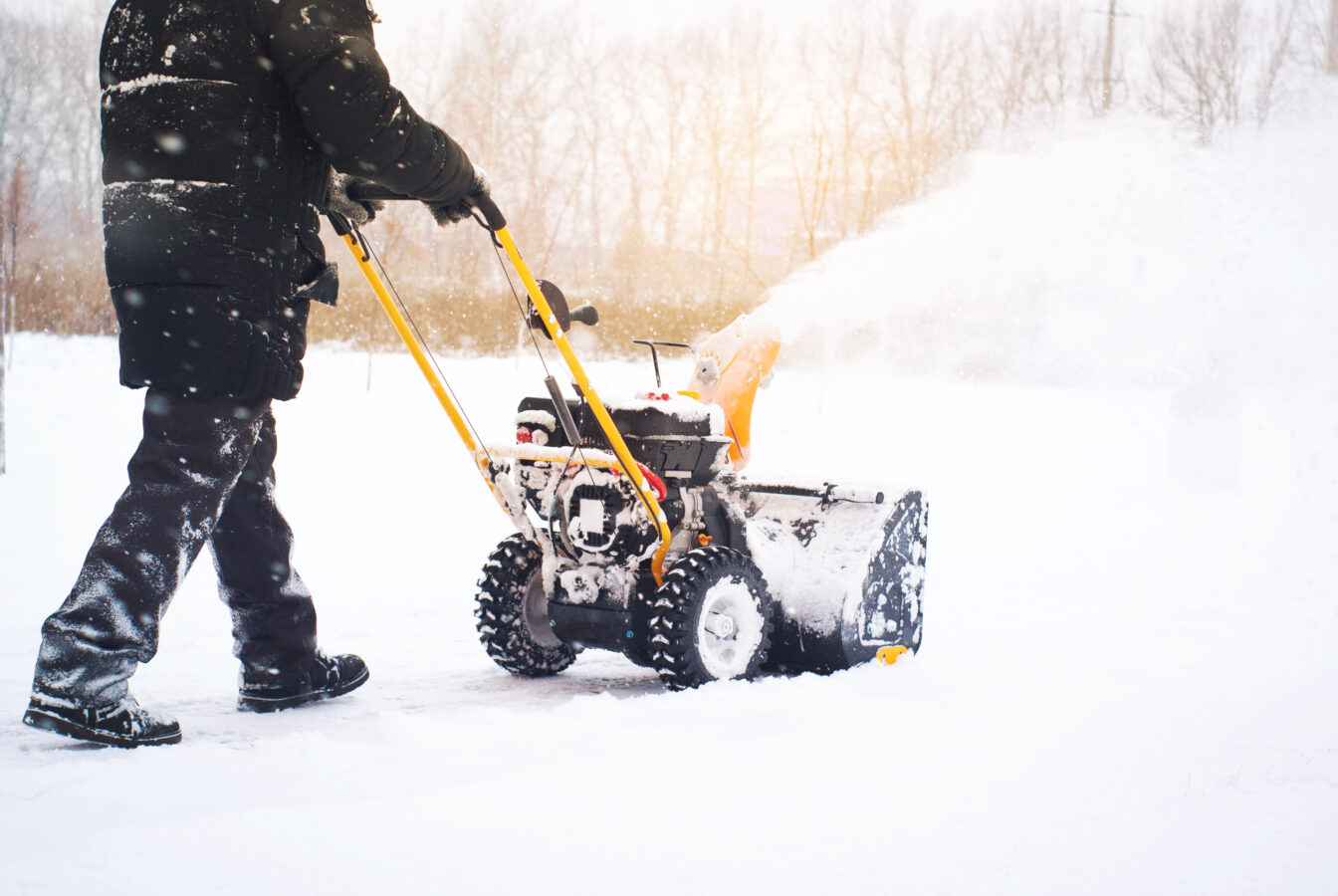 Person using snow blower in winter landscape with snow-covered ground and trees.