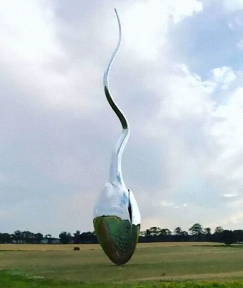 Large metallic spoon sculpture hanging upside down in a field under cloudy sky.