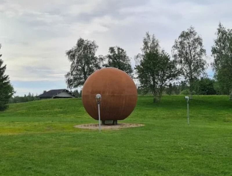 Large spherical sculpture on a grassy field with trees and cloudy sky in background.