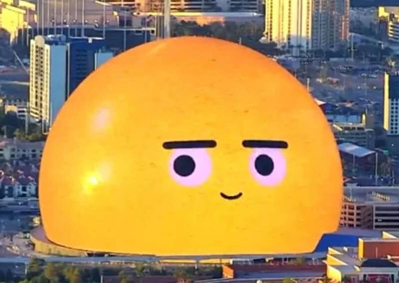 Large yellow dome with a cute, simple face on it in an urban cityscape.