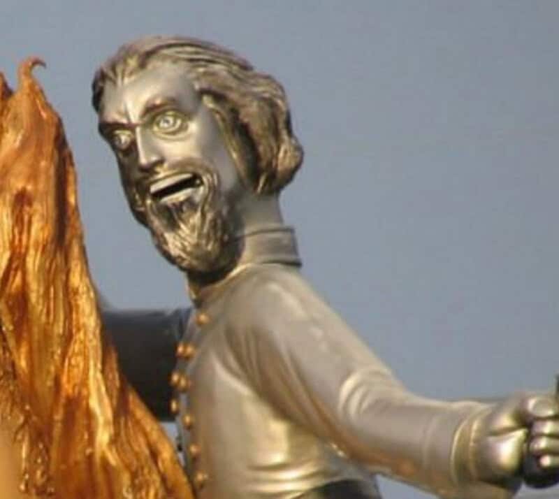 Silver statue of a man with a beard and long hair, smiling, with a wooden element on the left.
