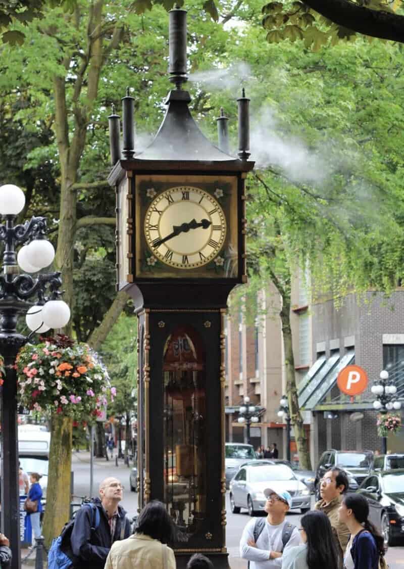 Vintage street clock emitting steam in a park setting with people walking and cars in the background.