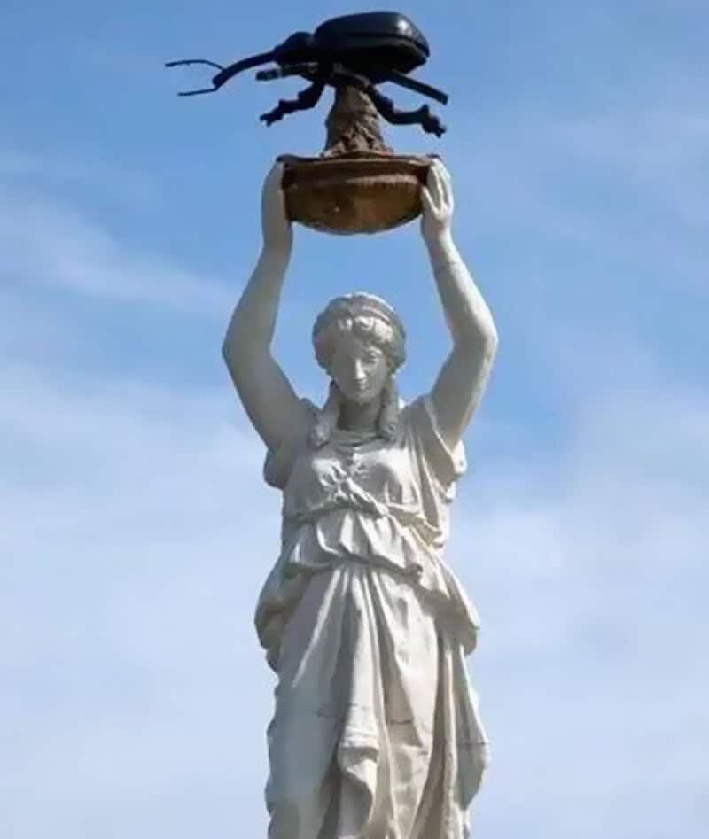 A woman holding a large beetle sculpture above her head on a statue.