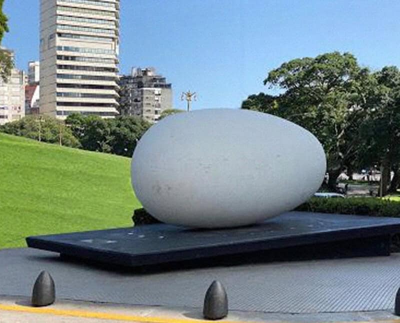 Large white egg sculpture on black platform in urban park setting.