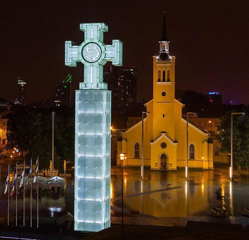 Illuminated cross and yellow church at night in a city square.