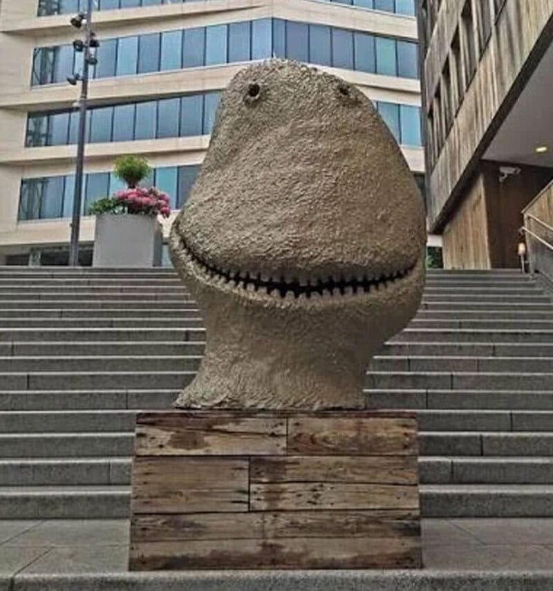 Large stone sculpture of a smiling dinosaur head on city stairs.