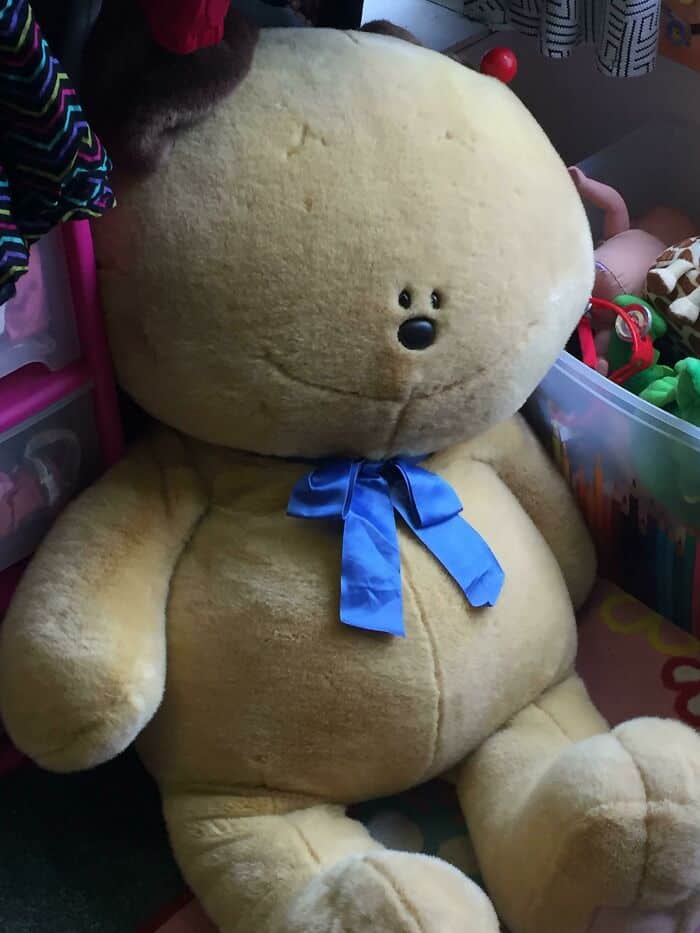 Soft plush teddy bear with a blue ribbon around its neck, sitting among toys and colorful objects.