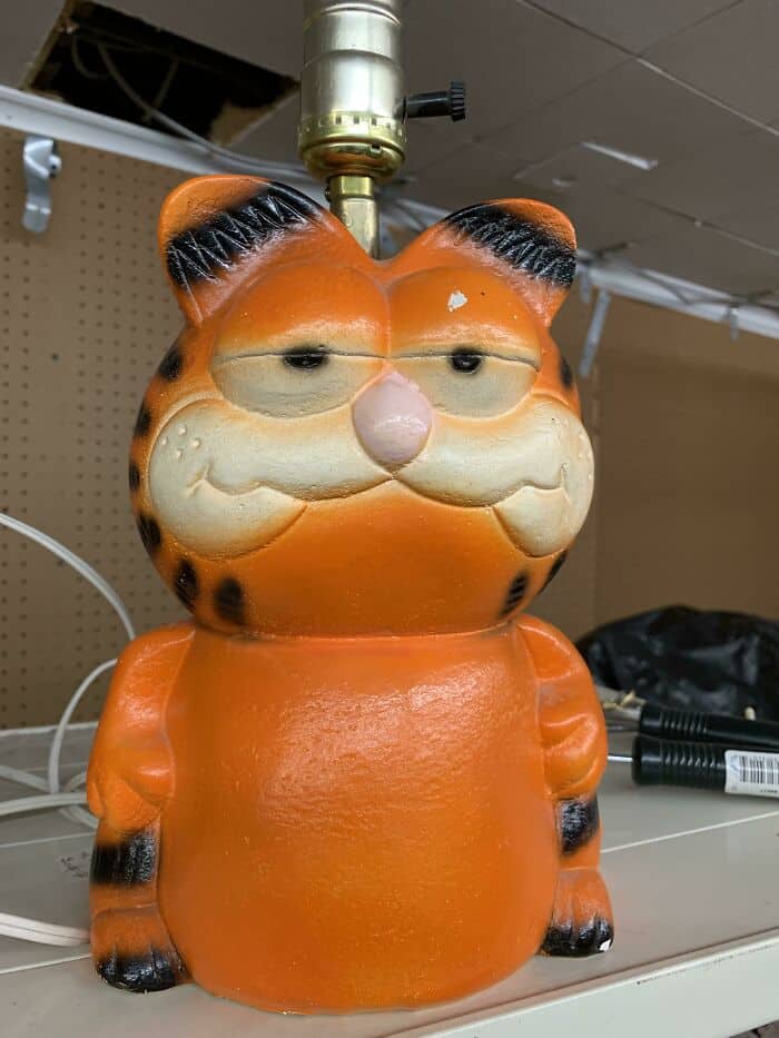 Orange tiger-shaped lamp with sleepy eyes and a pink nose on a shelf.