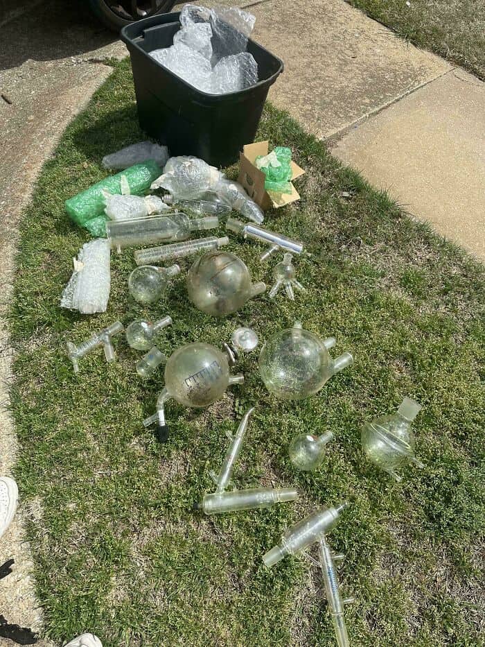 Broken glass bottles and containers on grass near trash bin.