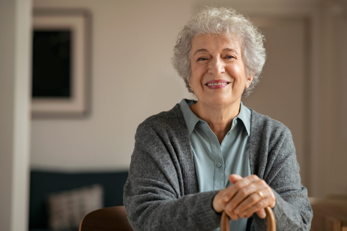 Smiling elderly woman with gray hair holding a cane in a cozy home.