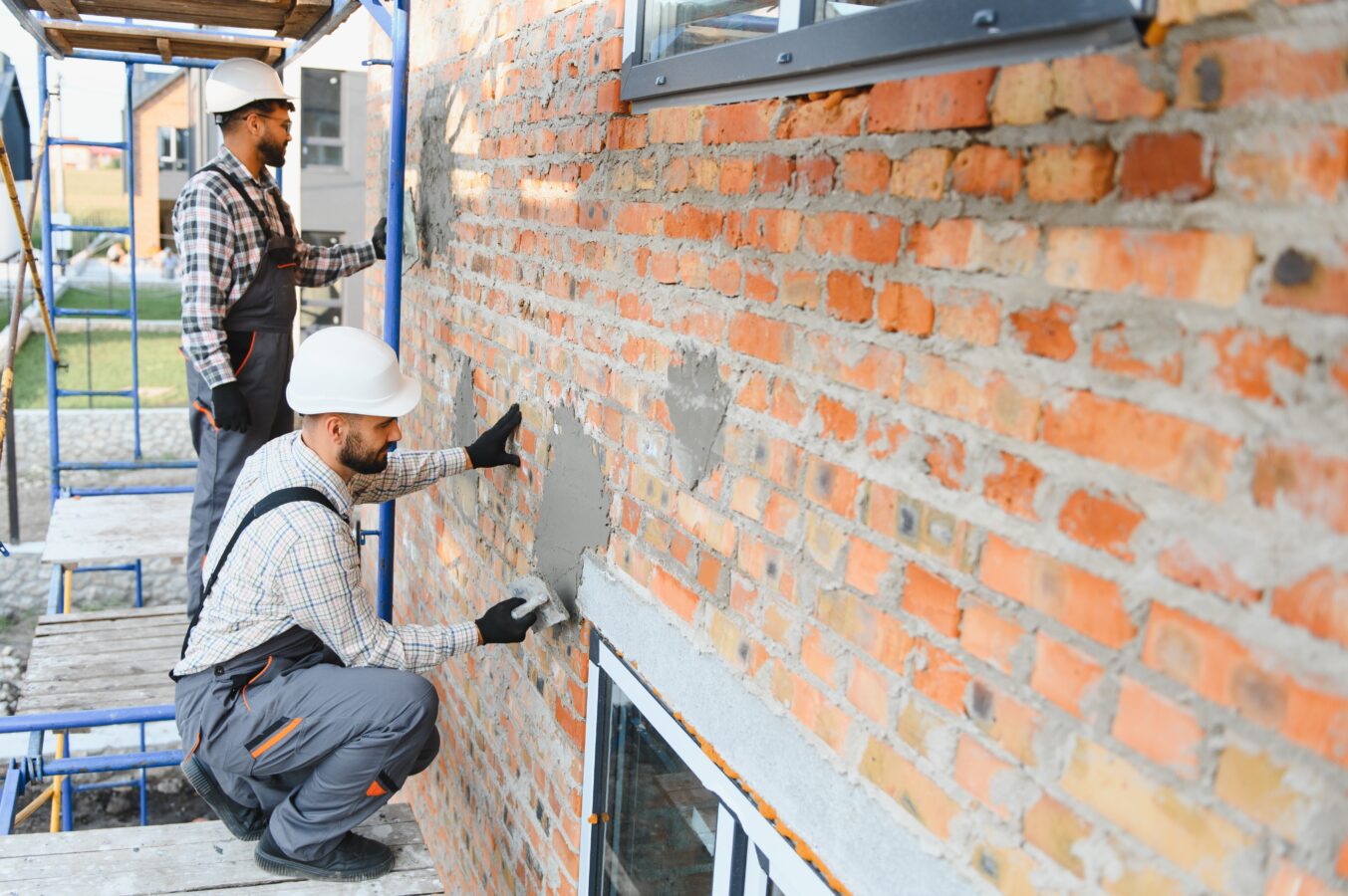 Two construction workers applying insulation to a brick wall outside.