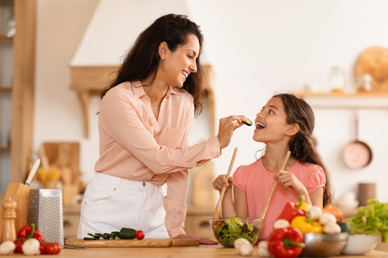 A woman feeding a girl in a kitchen with vegetables and cooking utensils.