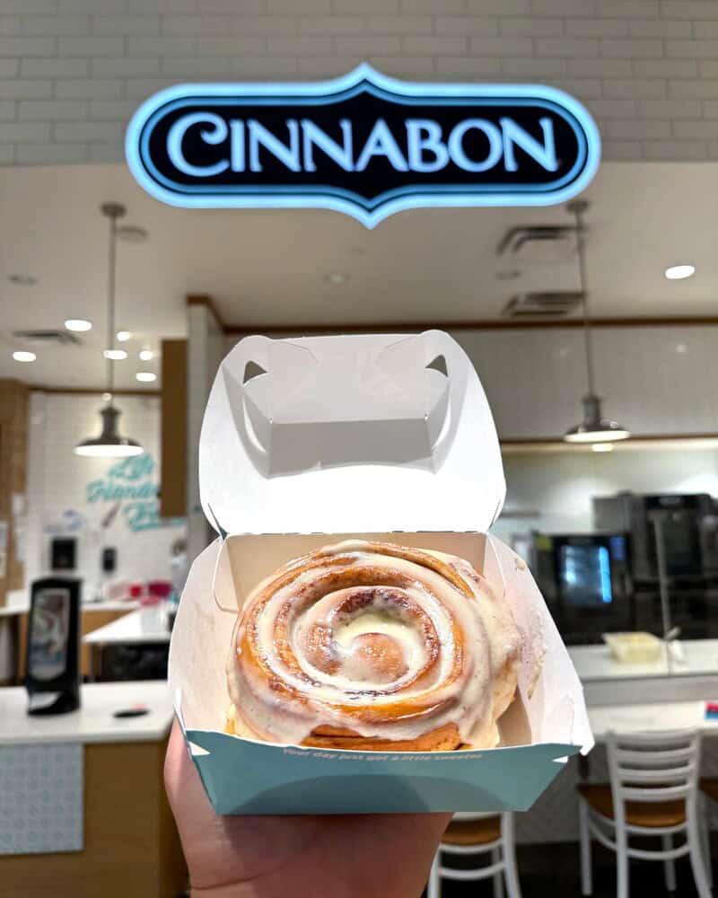 Cinnabon cinnamon roll in a box at the mall with a Cinnabon sign above.