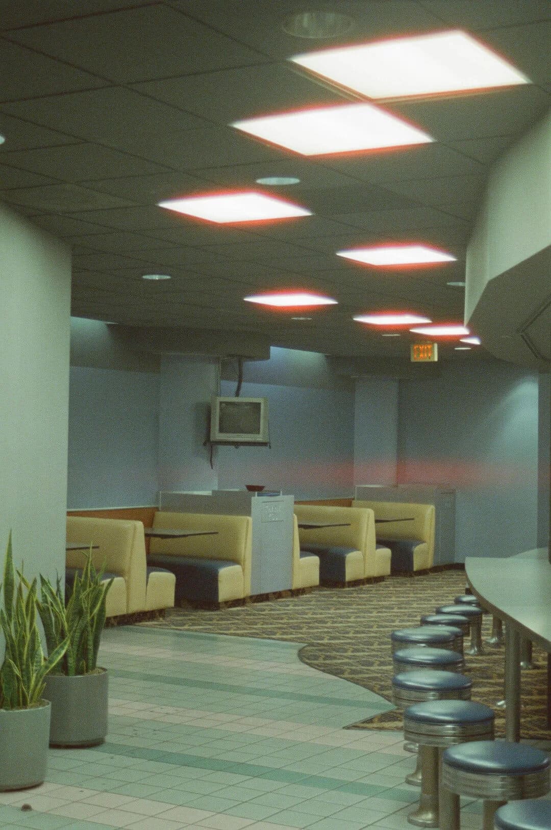 Empty retro diner with yellow booths and round bar stools, ceiling lights, and potted plants.