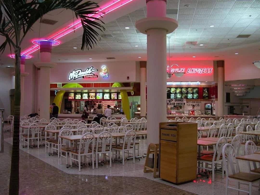 Food court with neon signs and white chairs, 90s design nostalgia.