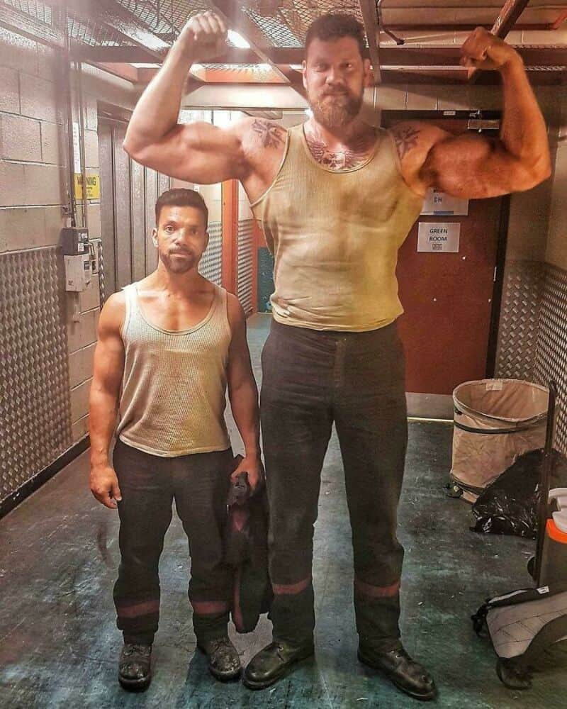 Two muscular men in tank tops flex their muscles in a gym.