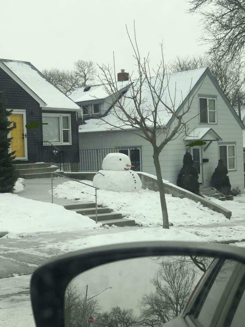 Snowman with a small snowball on top, in front of a house with snow, trees, and a driveway.