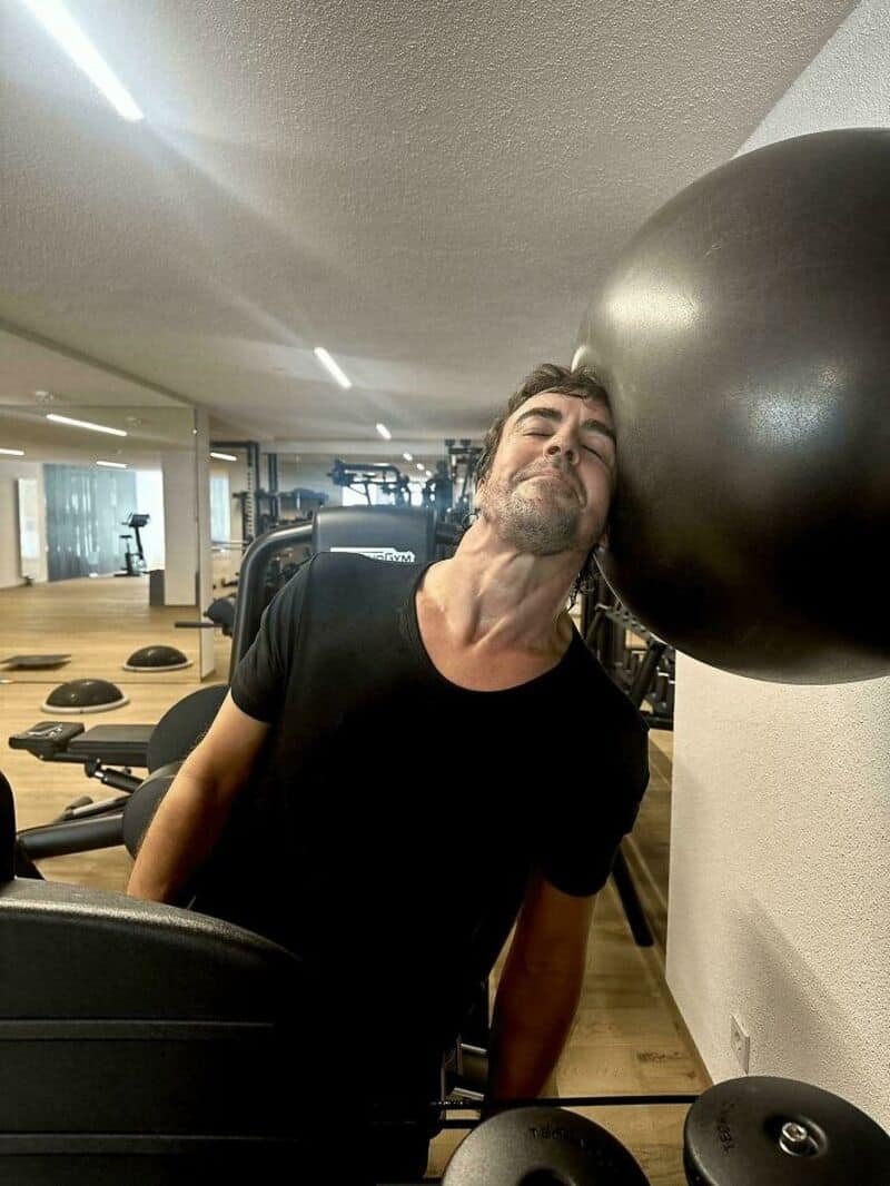 Man with beard leaning his head against a large black exercise ball in a gym.