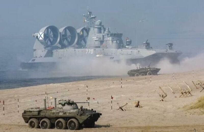 Large battleship with three large propellers and a smaller military vehicle on sandy beach.