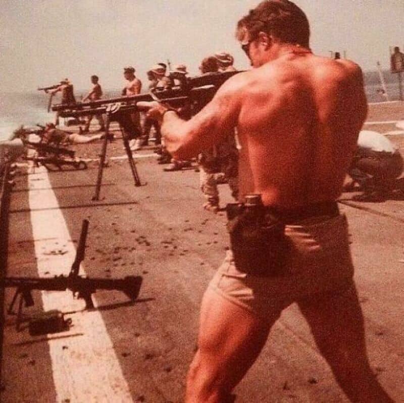 Man aiming a rifle on a military aircraft carrier with soldiers in the background.