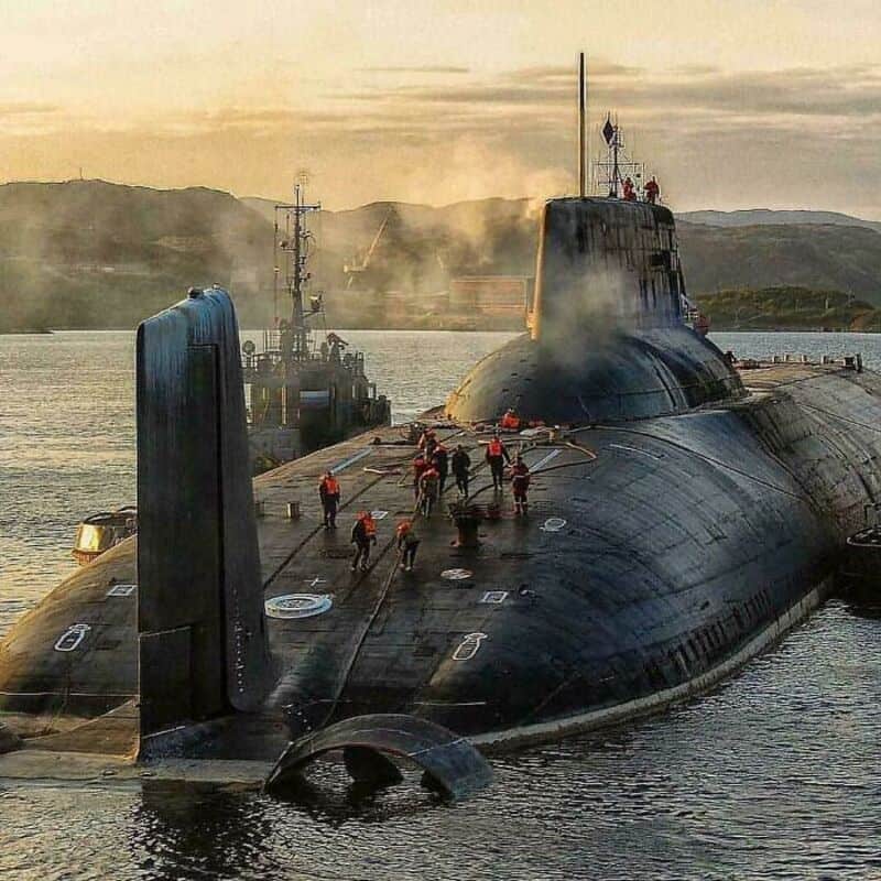 Submarine with crew on deck, large and imposing in water, mountains in background.