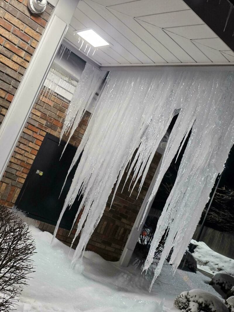 Large icicles hanging from a roof outside a building with brick wall and snow on the ground.