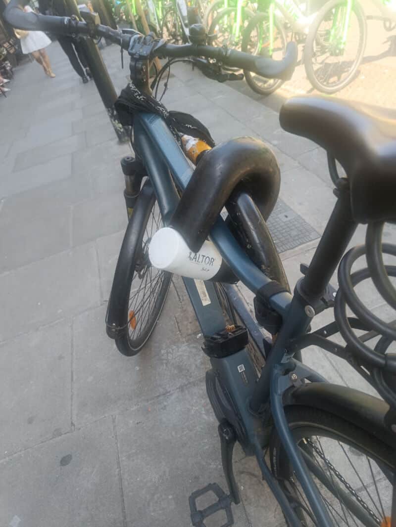 Close-up of a black e-bike with a white lock on the frame, parked on a sidewalk.