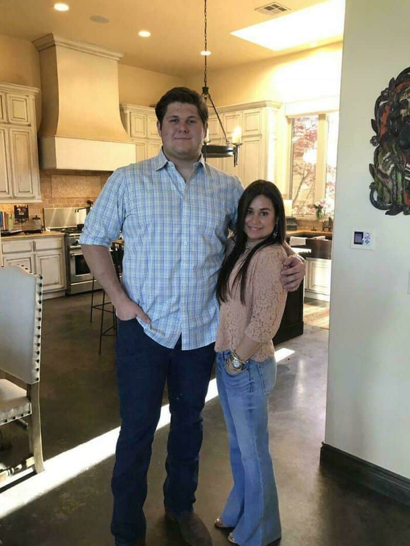 Two people standing in a kitchen, one tall man and one shorter woman, smiling.