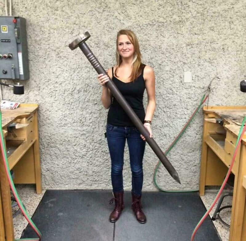 Woman holding a giant hammer, standing in workshop with wooden workbenches.