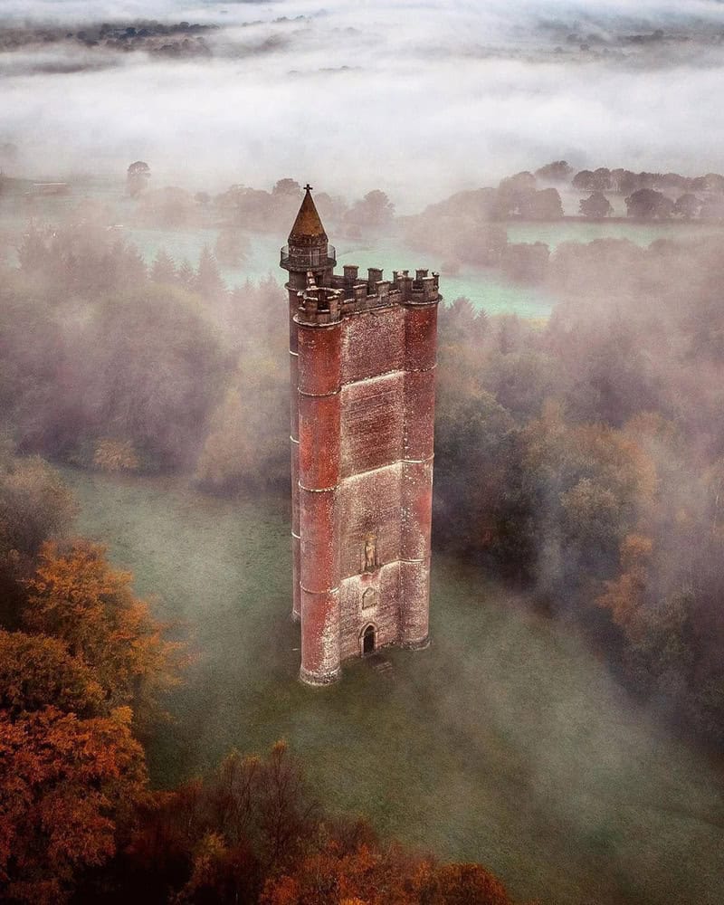 A tall, red brick tower with battlements, surrounded by fog and autumn trees.