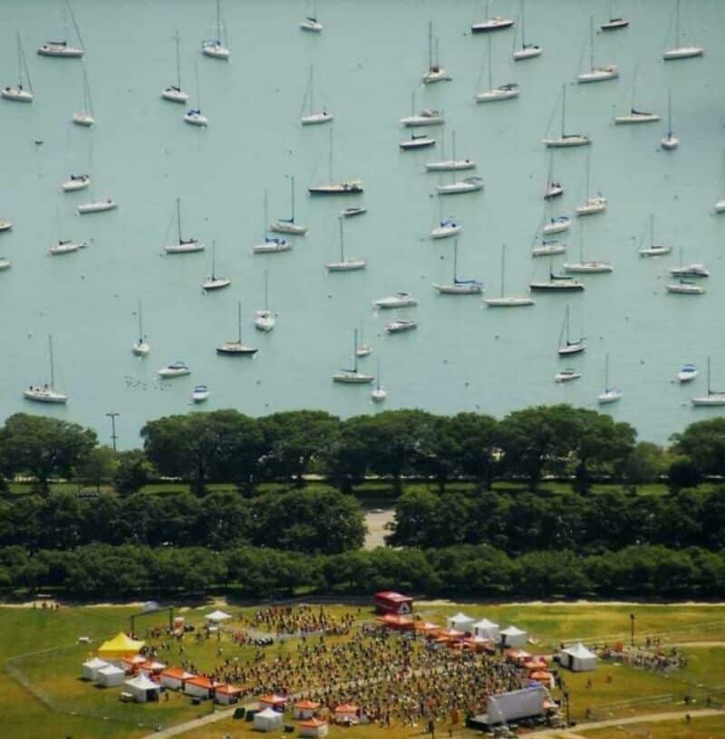 A surreal image showing boats floating above a park with a crowd and tents below.