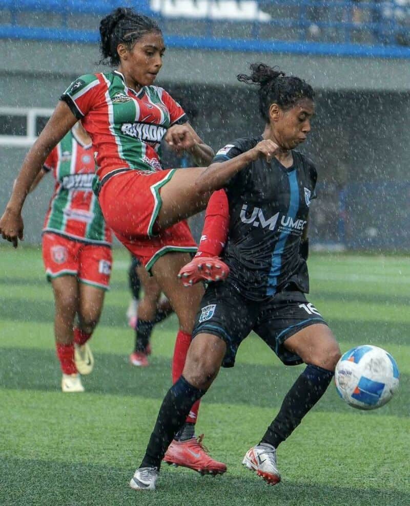 Two female soccer players competing in rain, one in red and green, the other in black, fighting for球.
