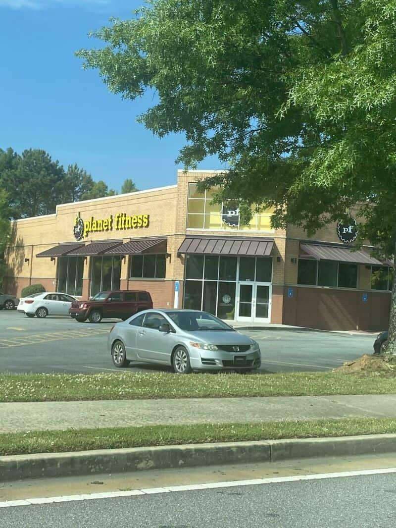 Planet Fitness gym in a shopping center with parked cars and green trees.