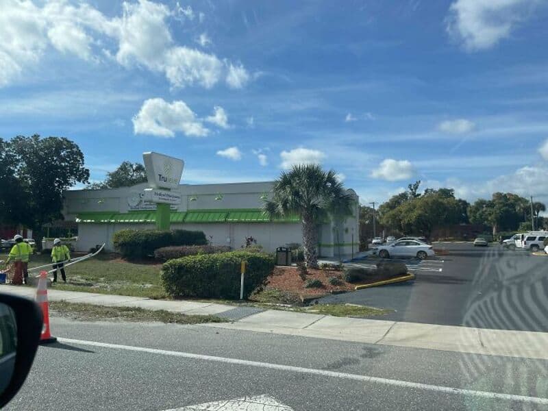 A building with a green roof and a sign that says 'Tru' with a parking lot and palm trees.