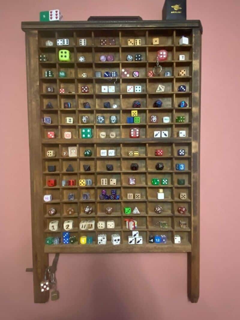 Wooden dice storage cabinet with various dice in compartments, against pink wall.